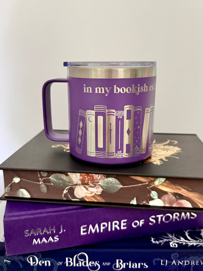 Purple stainless steel mug with 'in my bookish era' and books engraved around  it, sitting on a stack of black, purple and blue books.