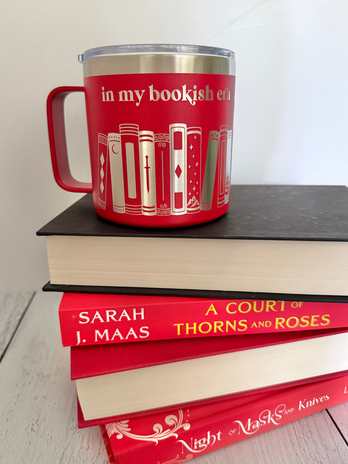 Red stainless steel mug with 'in my bookish era' and books engraved around it, set on top of a stack of red and black books.