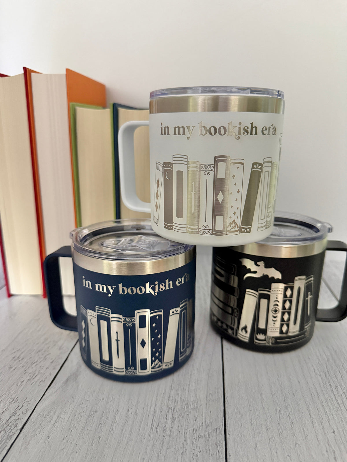 Three stainless steel mugs stacked in a pyramid with 'in my bookish era,' books and a dragon engraved around each, set on a white wood floor with books in the background.
