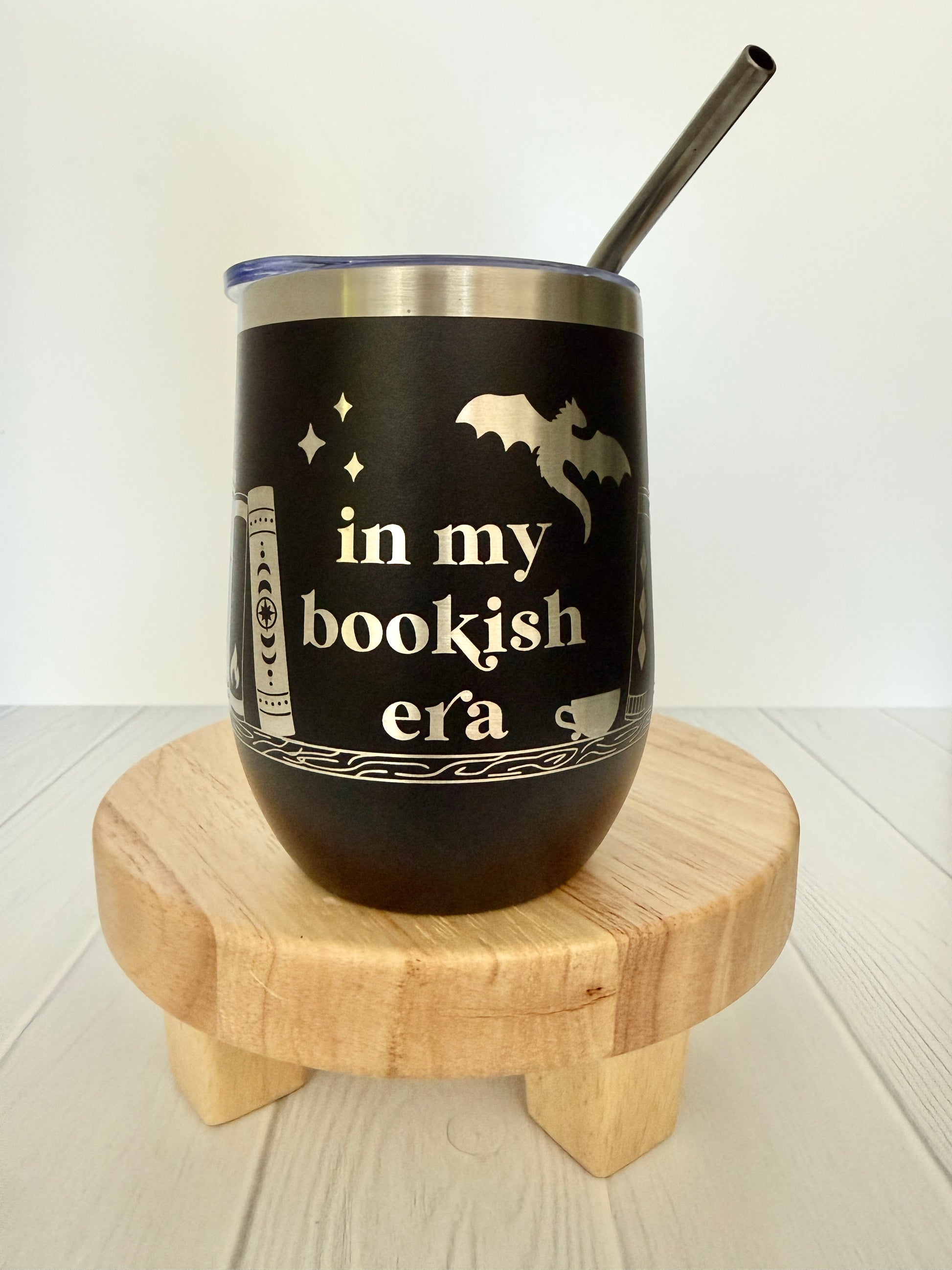 Black tumbler with 'in my bookish era' text and a straw on a wooden stand.