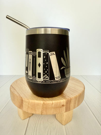 Black tumbler with book design on a wooden stand against a white background