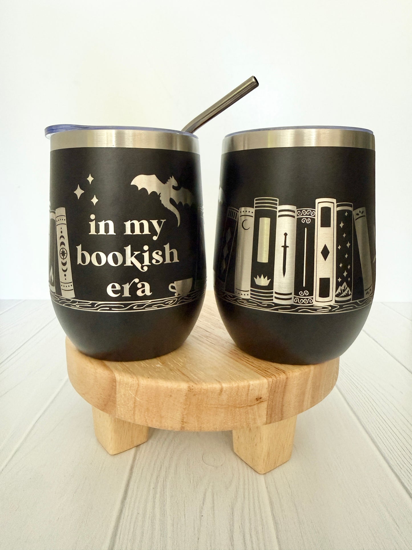 Two black tumblers with book-themed designs on a wooden stand.