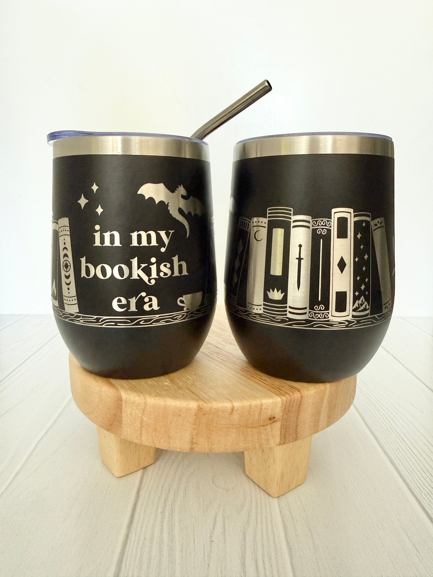 Two black tumblers with book-themed designs on a wooden stand.