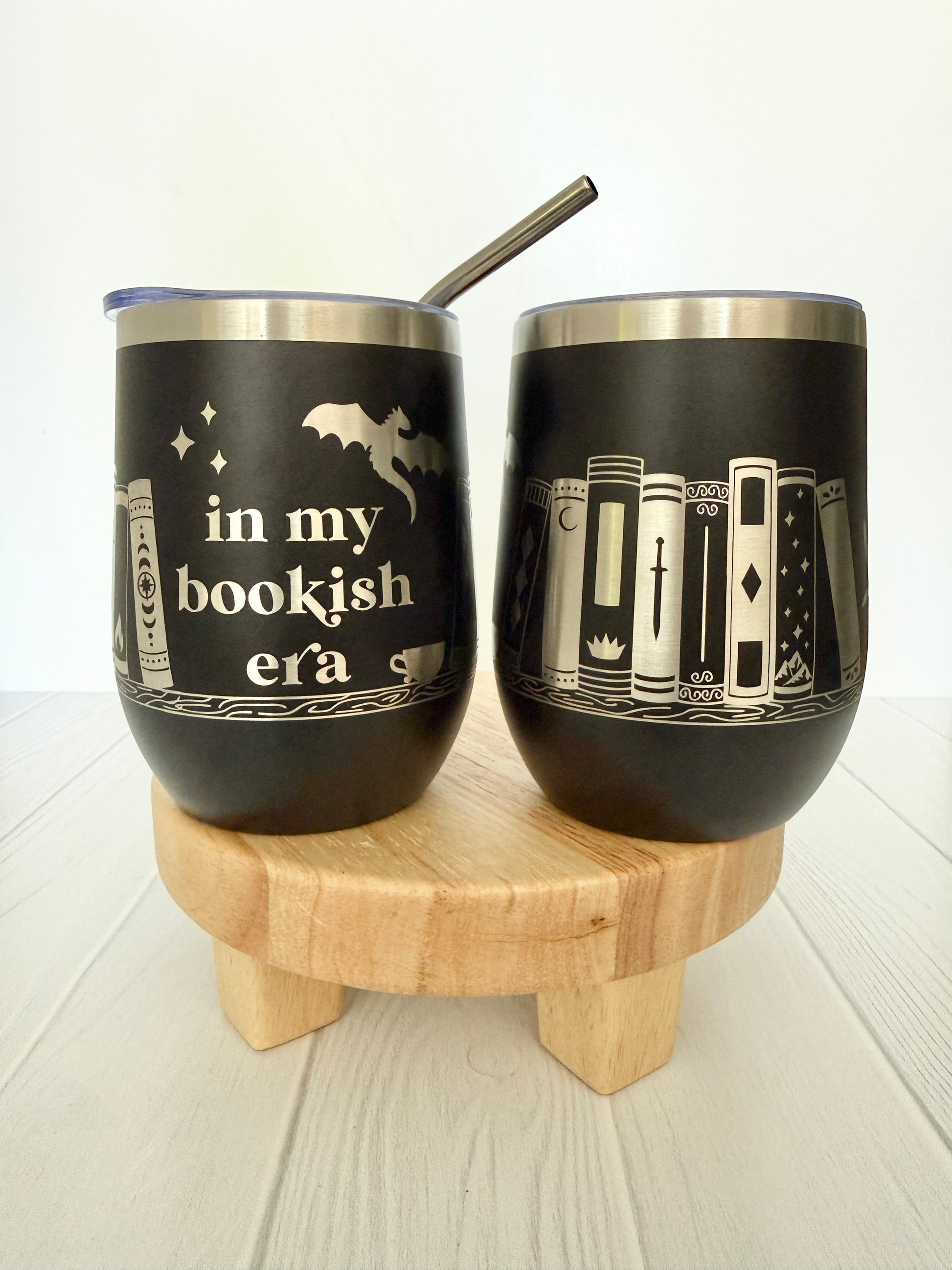 Two black tumblers with book-themed designs on a wooden stand.