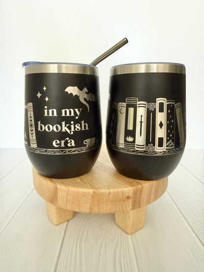 Two black tumblers with book-themed designs on a wooden stand.
