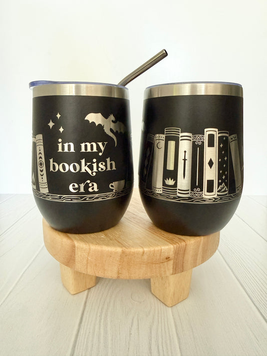 Two black tumblers with book-themed designs on a wooden stand.