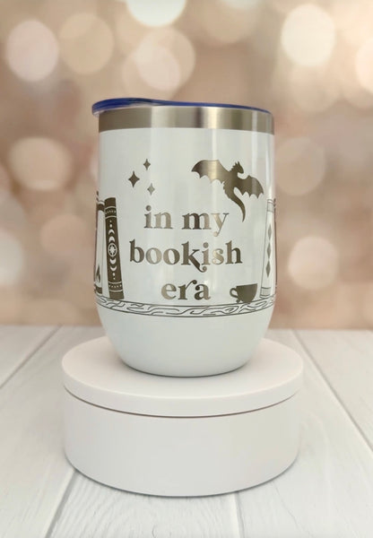 white wine tumbler engraved with "in my bookish era" and books on a book shelf and a dragon