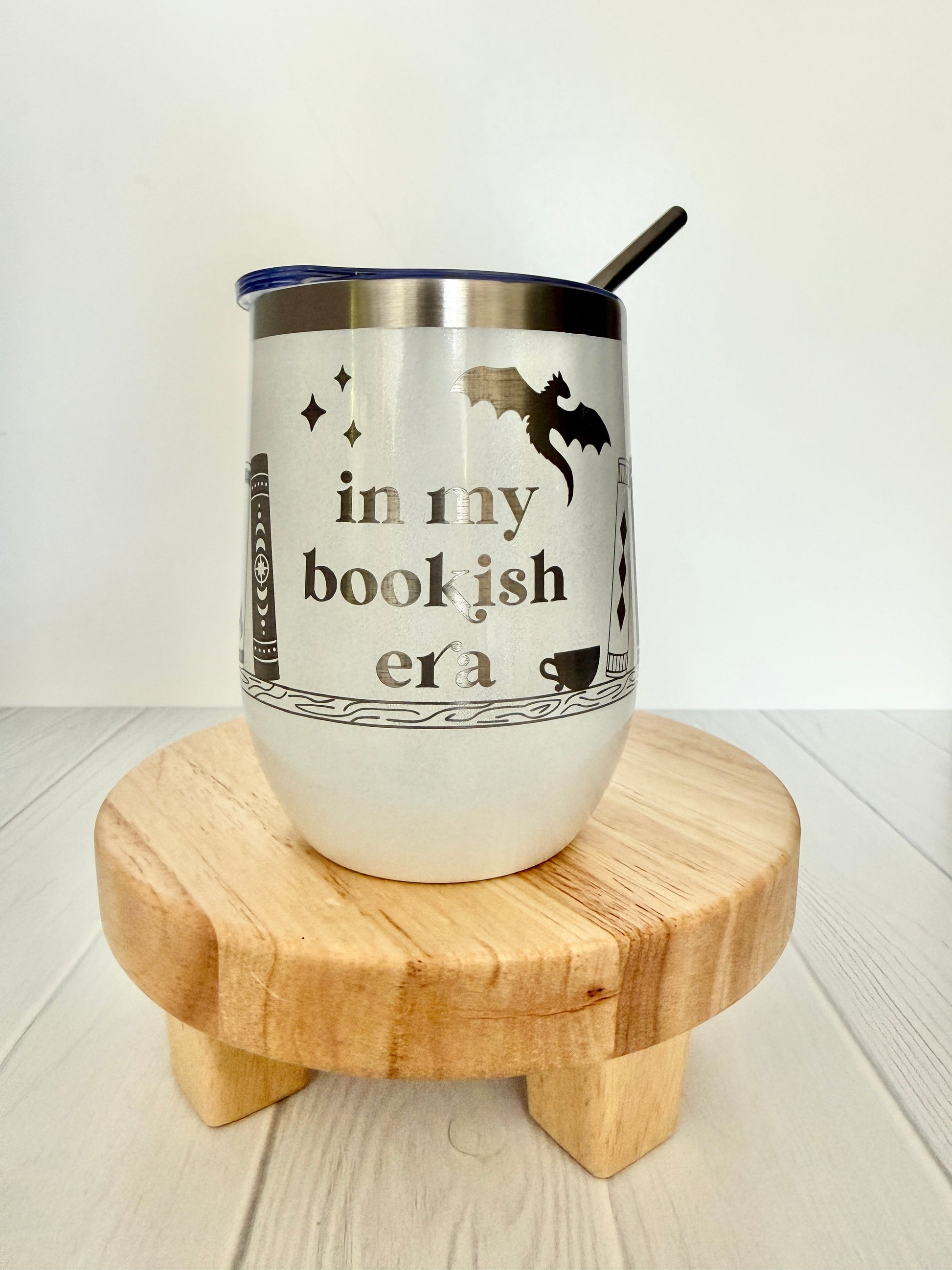 white wine tumbler engraved with "in my bookish era," books on a book shelf and a dragon with stars