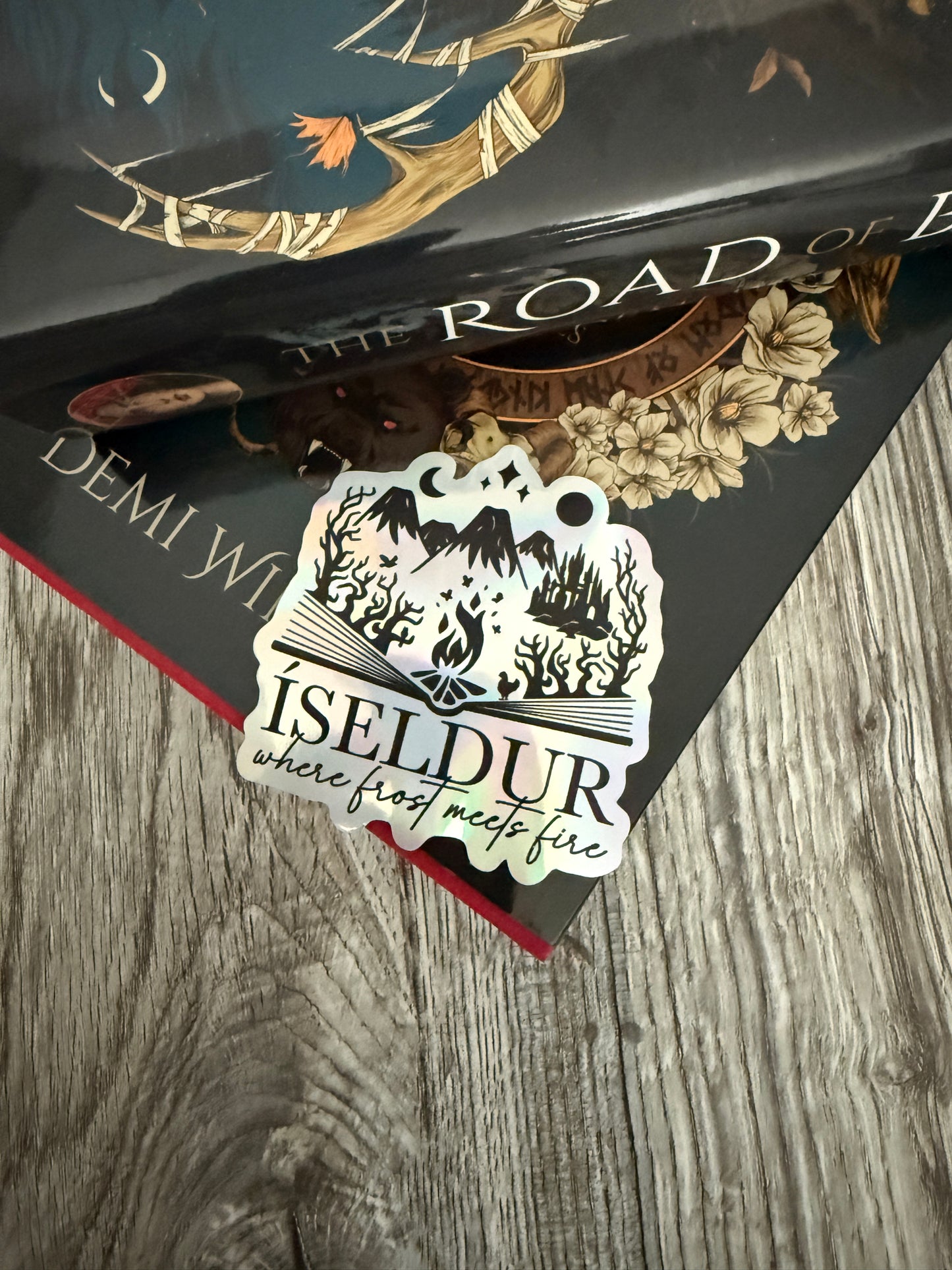 Holographic die cut sticker featuring an open book with fantasy elements and the words 'Iseldur: where frost meets fire' resting on The Road of Bones book by Demi Winters