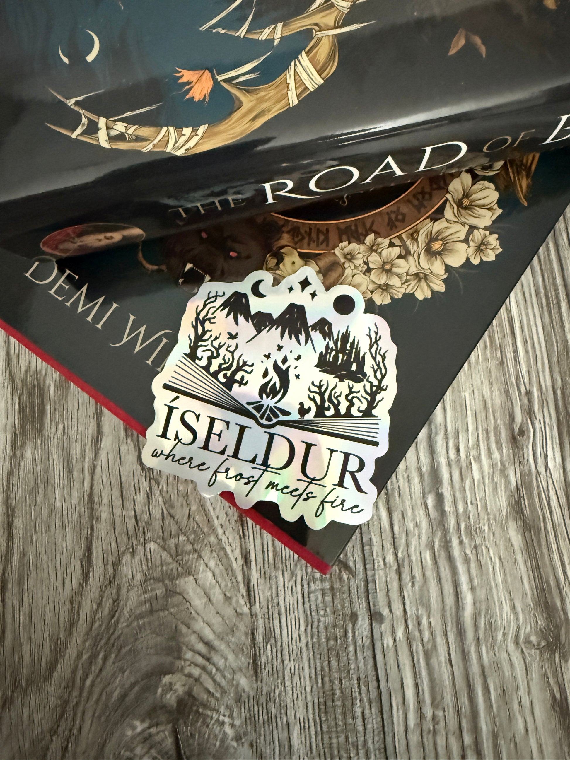 Holographic die cut sticker featuring an open book with fantasy elements and the words 'Iseldur: where frost meets fire' resting on The Road of Bones book by Demi Winters