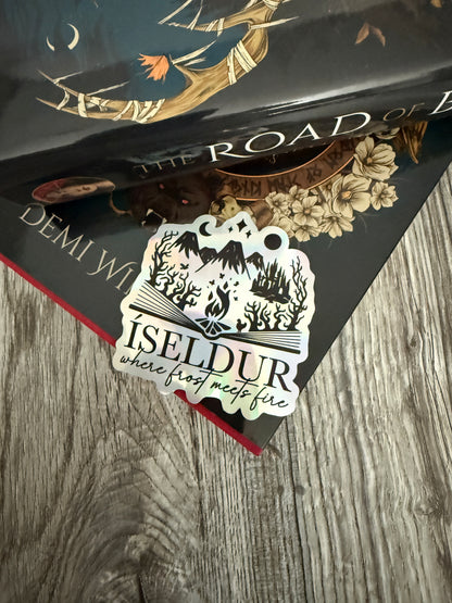 Holographic die cut sticker featuring an open book with fantasy elements and the words 'Iseldur: where frost meets fire' resting on The Road of Bones book by Demi Winters