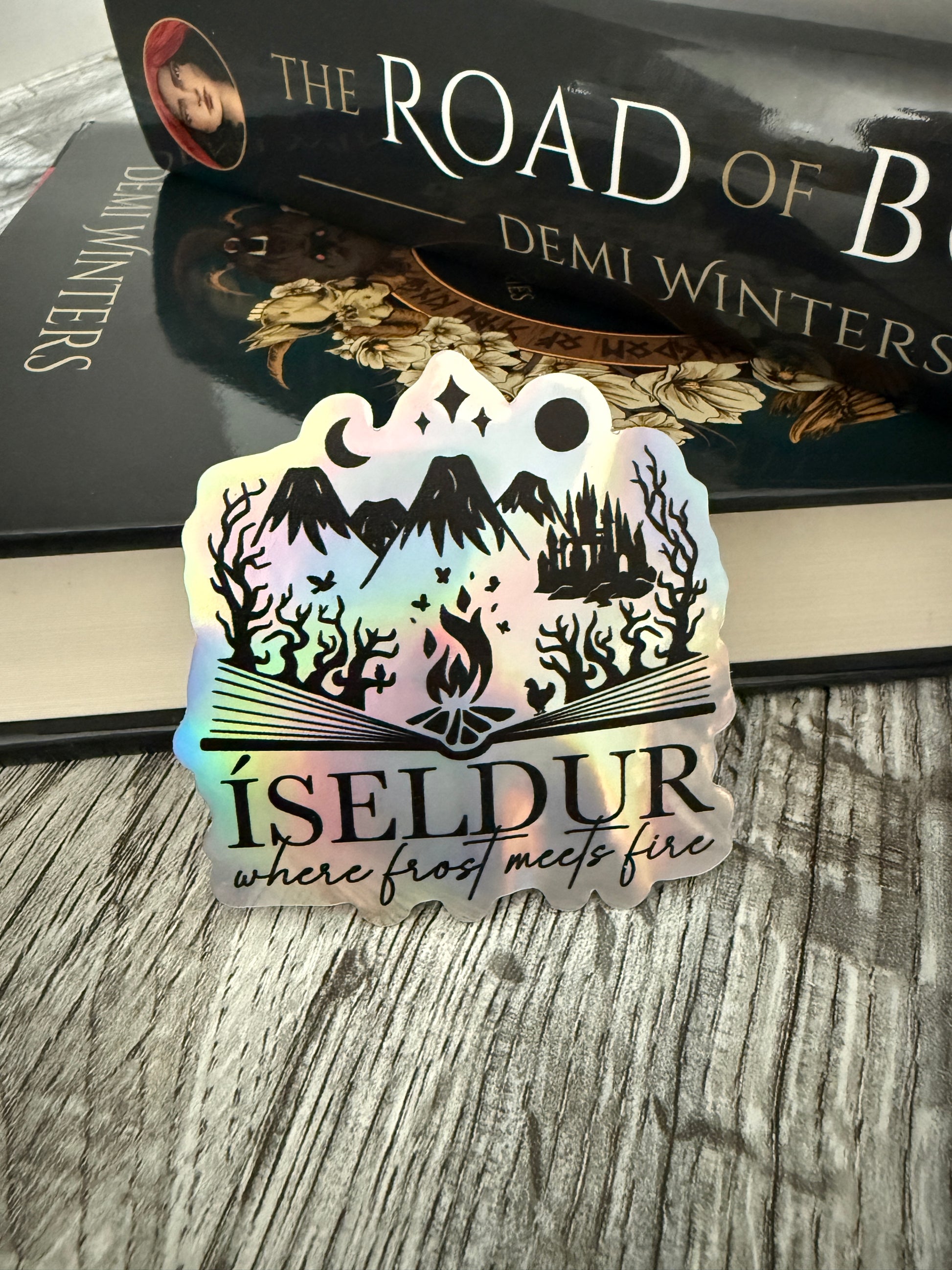 Holographic 'Isedur: where frost meets fire' sticker next to a book titled 'The Road of Bones' by Demi Winters on a wood surface.