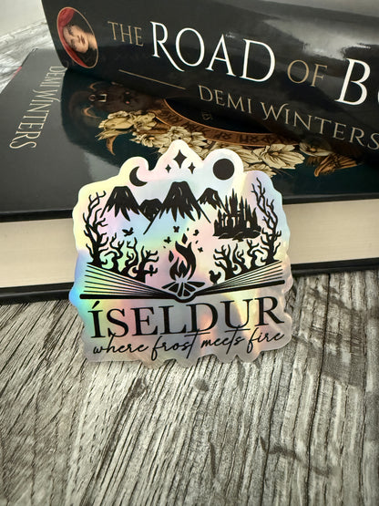Holographic 'Isedur: where frost meets fire' sticker next to a book titled 'The Road of Bones' by Demi Winters on a wood surface.