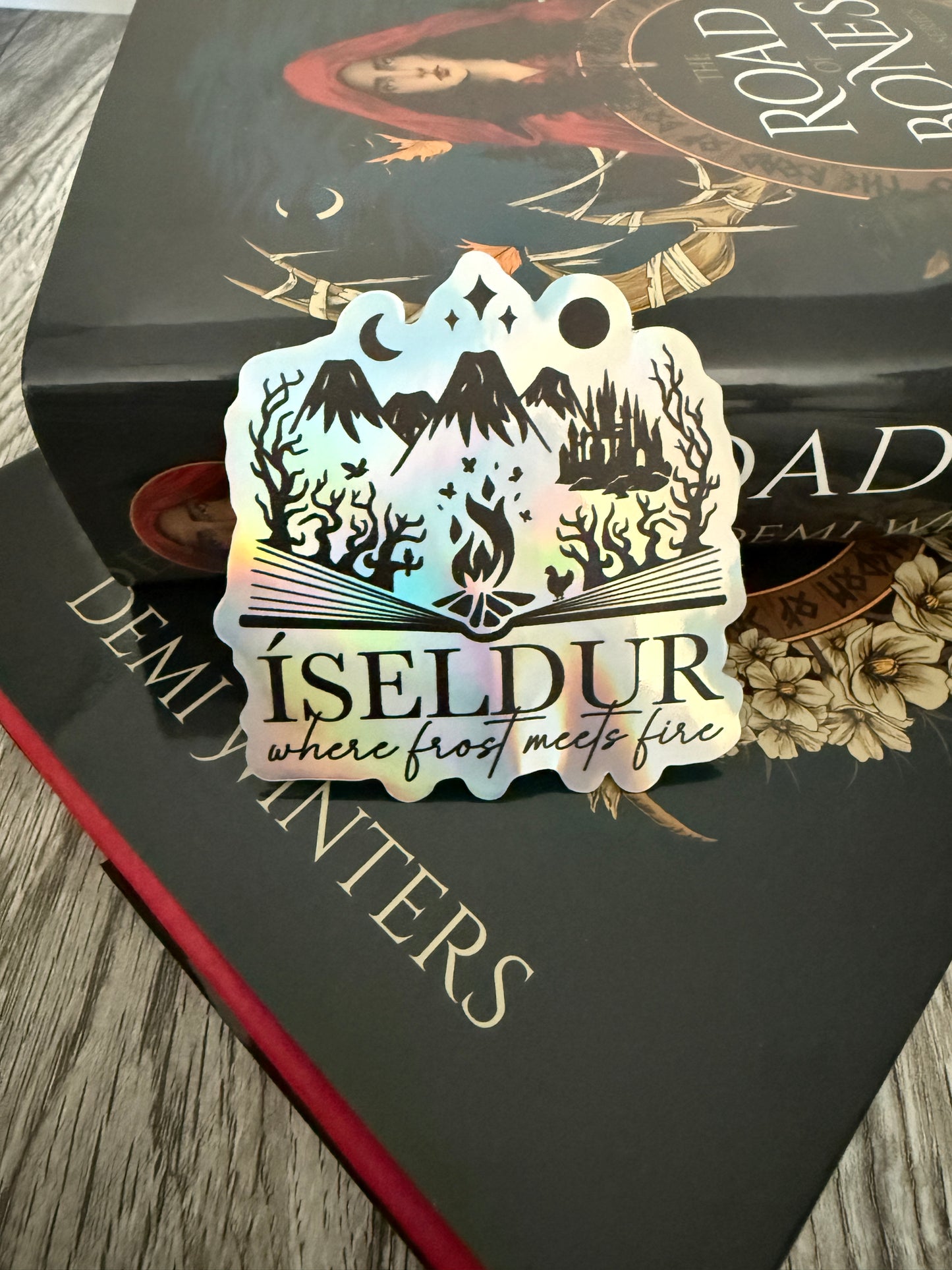 Road of Bones book by Demi Winters with a holographic sticker featuring an open book and the words 'Iseldur: where frost meets fire' on a wooden surface