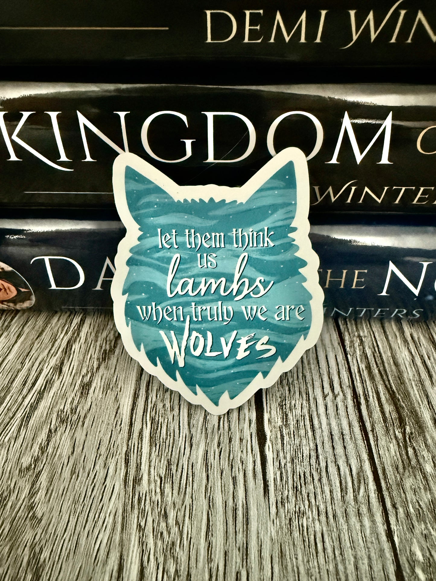 Sticker with wolf design and text 'let them think us lambs, when truly we are wolves' on a book stack. Inspired by Demi Winters' Ashen series.