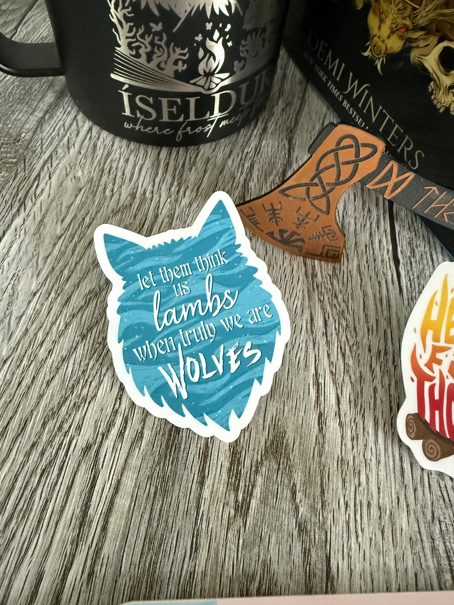 Sticker with wolf design and text 'let them think us lambs, when truly we are wolves' inspired by Demi Winters' Road of Bones book, on a wooden surface with books and a mug.