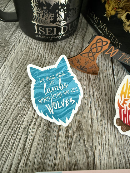 Sticker with wolf design and text 'let them think us lambs, when truly we are wolves' inspired by Demi Winters' Road of Bones book, on a wooden surface with books and a mug.