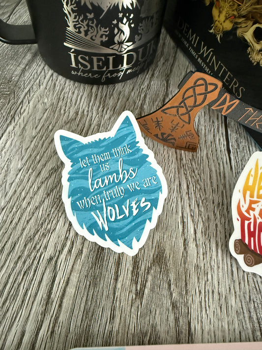Sticker with wolf design and text 'let them think us lambs, when truly we are wolves' inspired by Demi Winters' Road of Bones book, on a wooden surface with books and a mug.