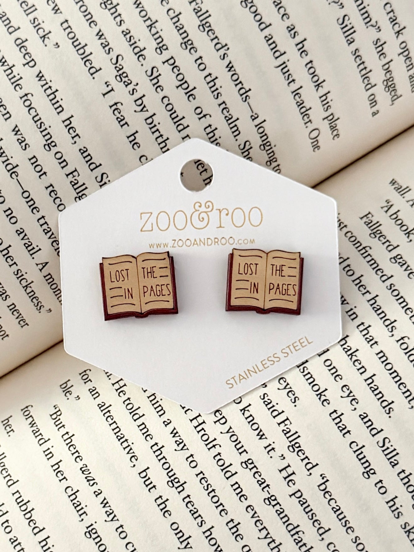 A pair of book-shaped stud earrings displayed on a white zoo&roo branded card, placed on top of open book pages. The book earrings are engraved with 'LOST IN THE PAGES.'