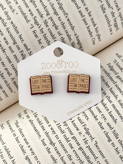 A pair of book-shaped stud earrings displayed on a white zoo&roo branded card, placed on top of open book pages. The book earrings are engraved with 'LOST IN THE PAGES.'