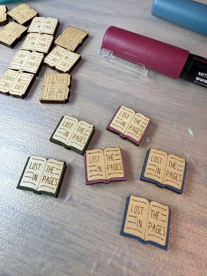 Wood stud earrings in the shape of books with 'lost in the pages' engraved on them, being hand painted