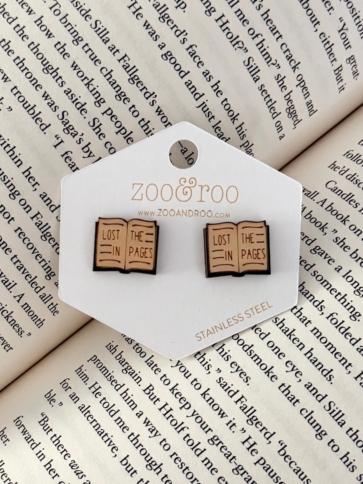 A pair of book-shaped stud earrings displayed on a white zoo&roo branded card, placed on top of open book pages. The book earrings are engraved with 'LOST IN THE PAGES.'