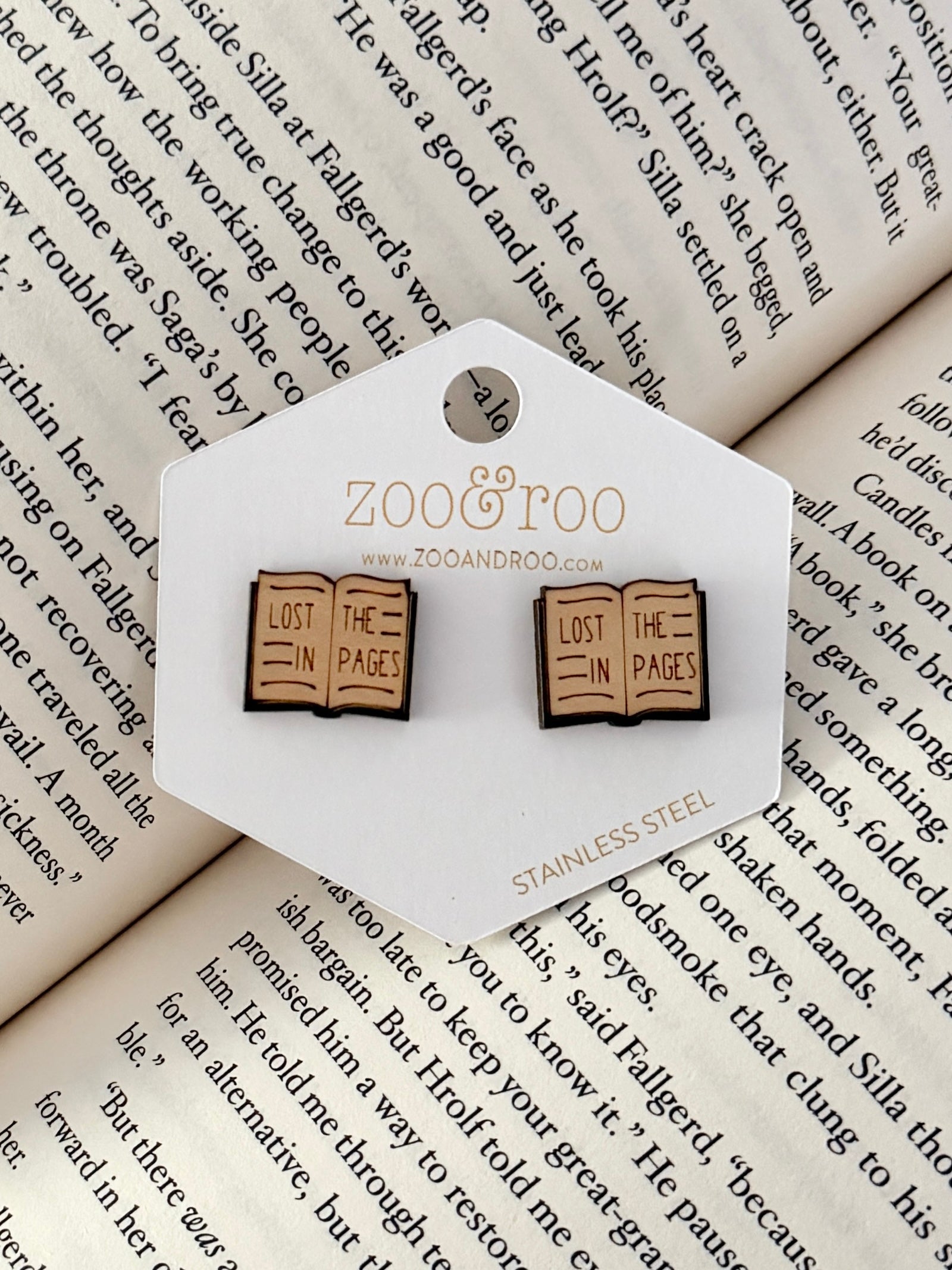 A pair of book-shaped stud earrings displayed on a white zoo&roo branded card, placed on top of open book pages. The book earrings are engraved with 'LOST IN THE PAGES.'