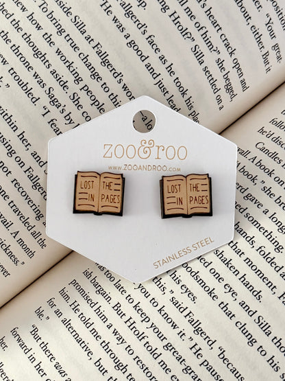 A pair of book-shaped stud earrings displayed on a white zoo&roo branded card, placed on top of open book pages. The book earrings are engraved with 'LOST IN THE PAGES.'