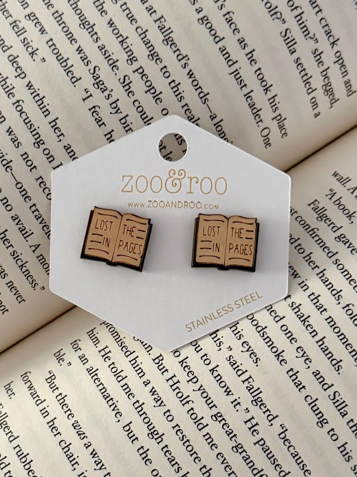 A pair of book-shaped stud earrings displayed on a white zoo&roo branded card, placed on top of open book pages. The book earrings are engraved with 'LOST IN THE PAGES.'