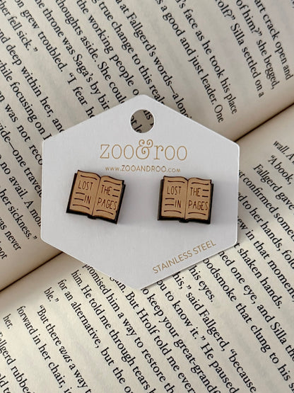 A pair of book-shaped stud earrings displayed on a white zoo&roo branded card, placed on top of open book pages. The book earrings are engraved with 'LOST IN THE PAGES.'