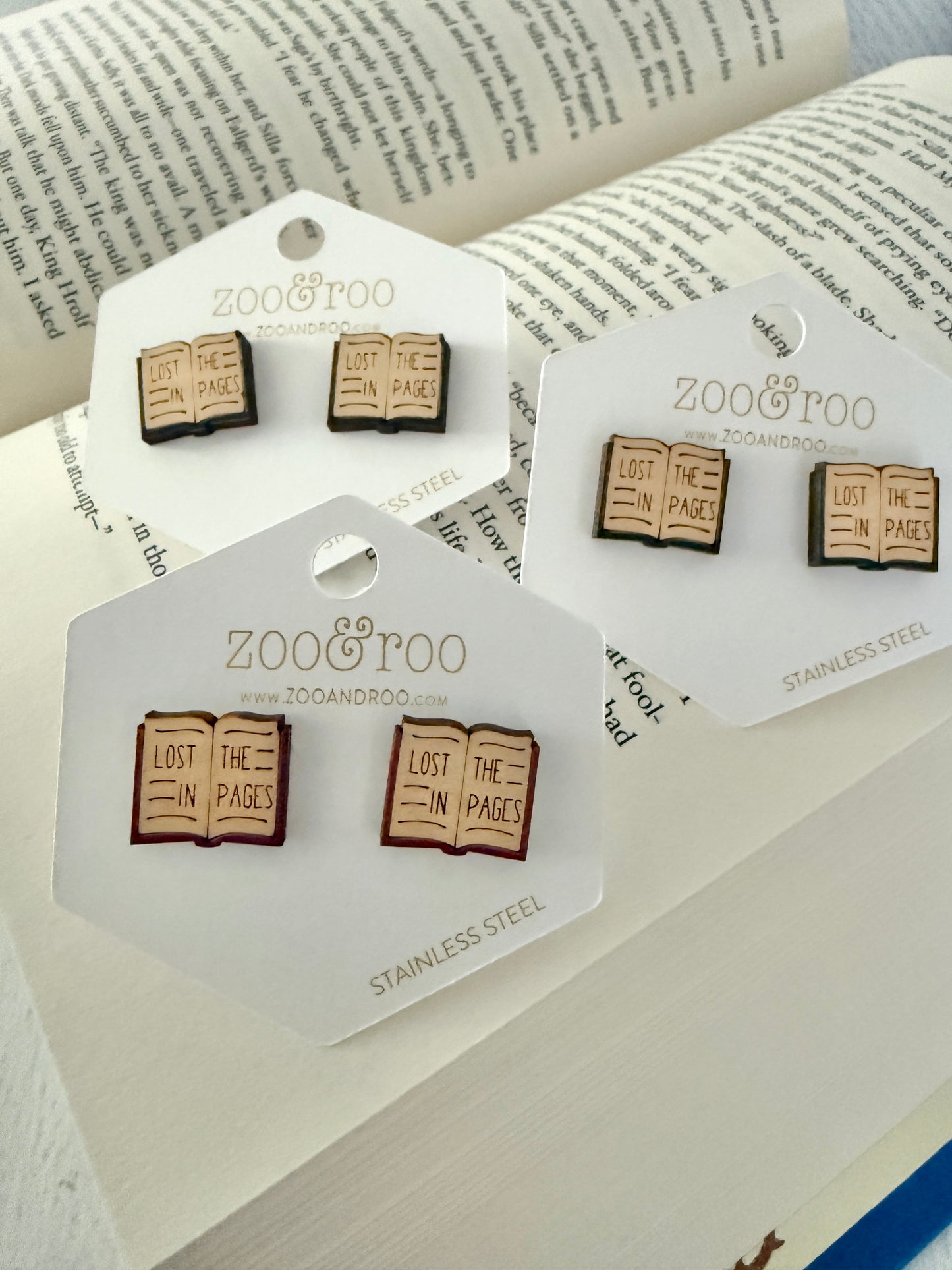 Three pairs of Zoo & Too book-themed stud earrings displayed on white branded cards, arranged on an open book background. The earrings depict open books with the phrase 'LOST IN THE PAGES'.
