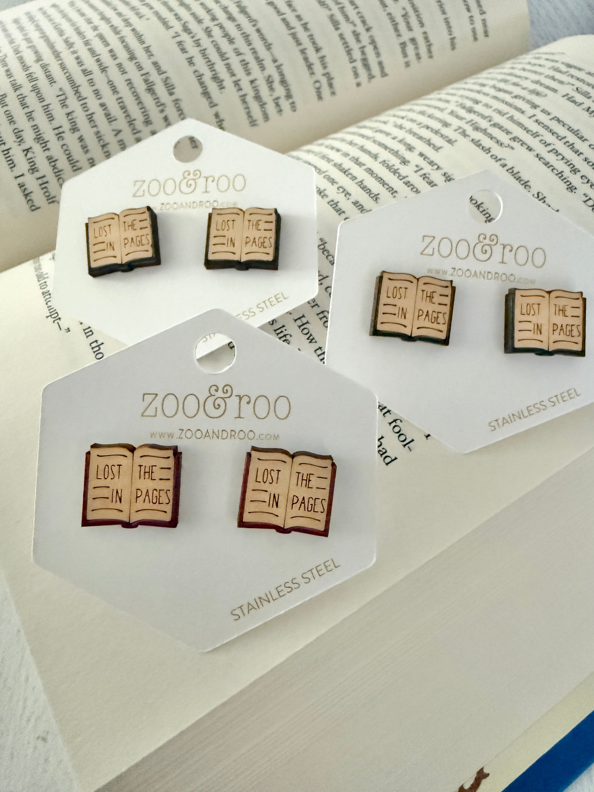 Three pairs of Zoo & Too book-themed stud earrings displayed on white branded cards, arranged on an open book background. The earrings depict open books with the phrase 'LOST IN THE PAGES'.
