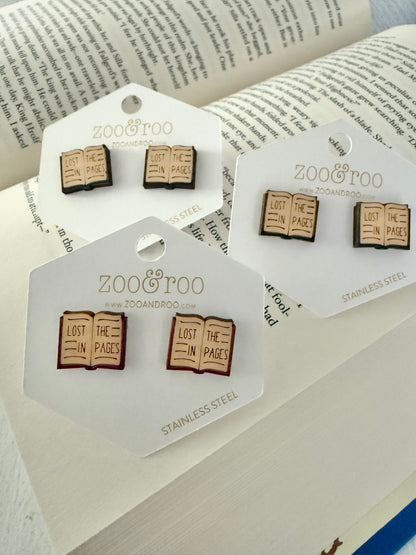 Three pairs of Zoo & Too book-themed stud earrings displayed on white branded cards, arranged on an open book background. The earrings depict open books with the phrase 'LOST IN THE PAGES'.