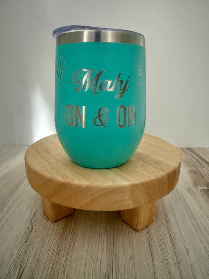 A turquoise insulated tumbler with a silver stainless steel rim and lid, featuring the engraved text 'Mahj ON & ON,' displayed on a small round wooden stand over a light wood floor against a neutral background.
