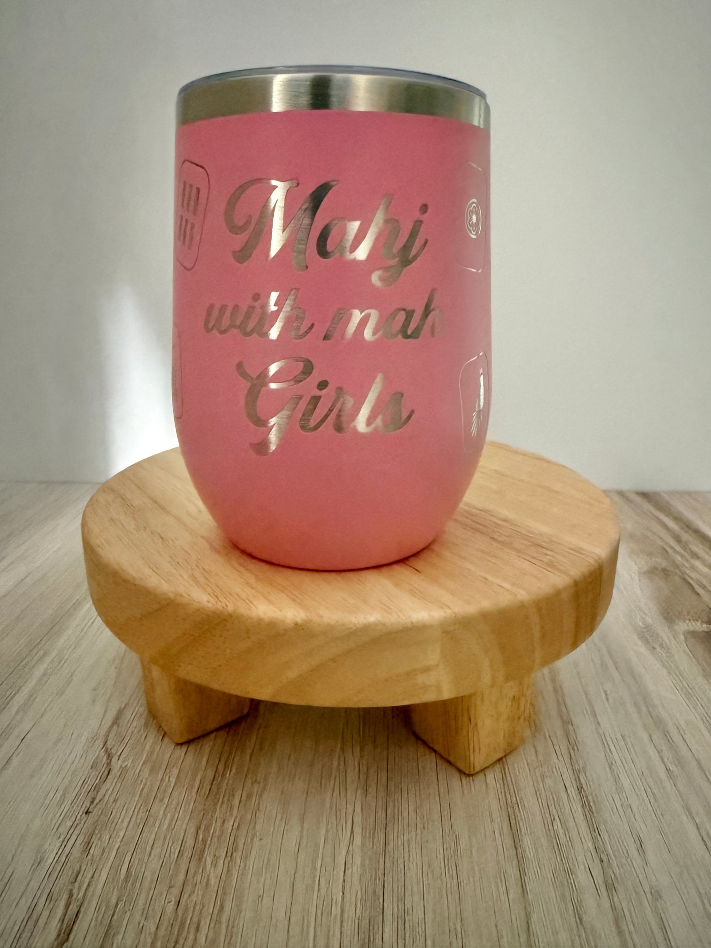 A pink insulated mahjong wine tumbler with a stainless steel rim, featuring the engraved text 'Mahj with mah Girls,' displayed on a small round wooden stand over a light wood surface.