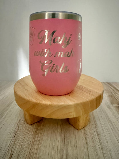 A pink insulated mahjong wine tumbler with a stainless steel rim, featuring the engraved text 'Mahj with mah Girls,' displayed on a small round wooden stand over a light wood surface.