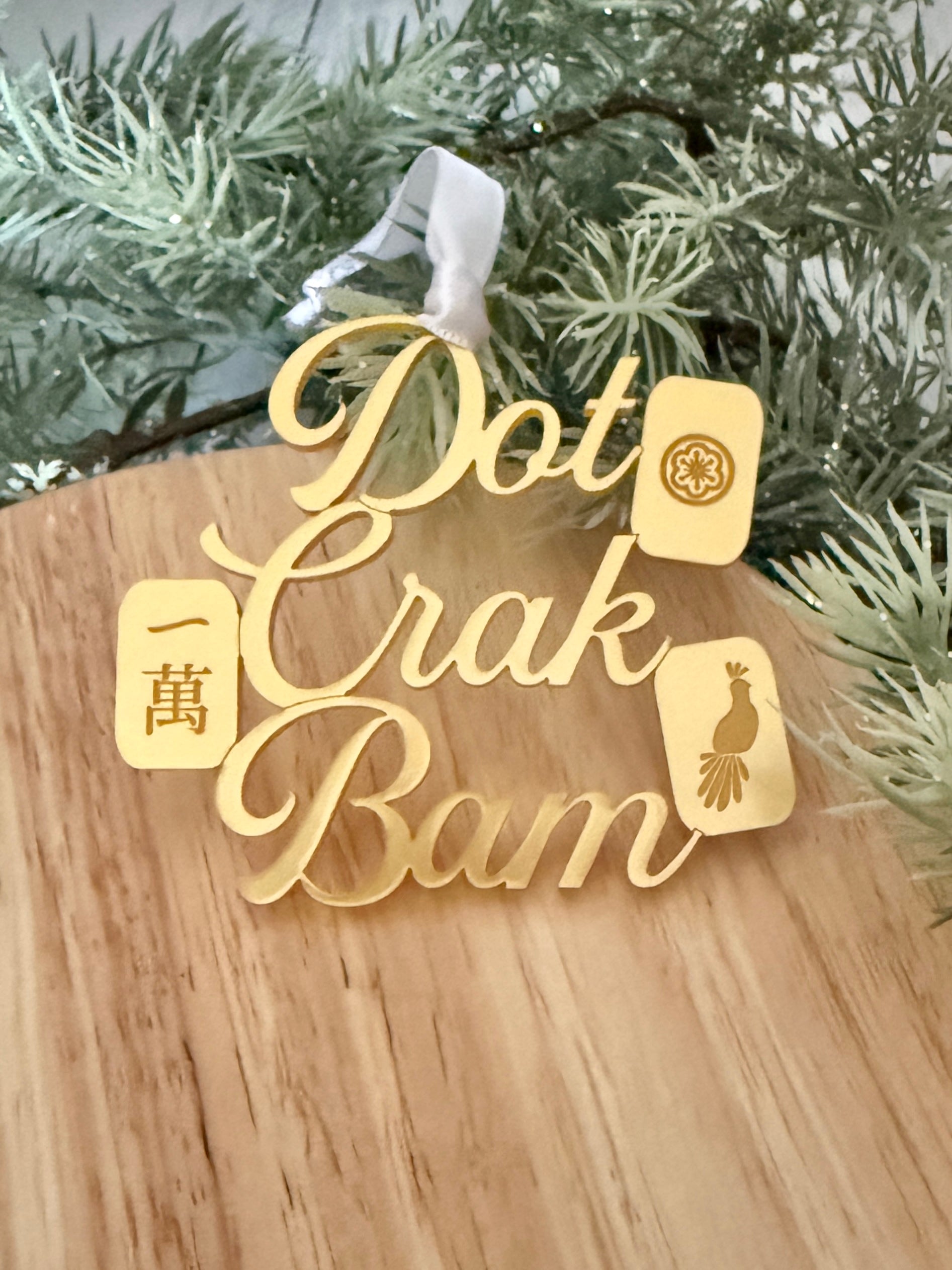 A gold metallic mahjong Christmas ornament spelling out 'Dot Crak Bam' with traditional mahjong tiles, hanging from a white ribbon on a wooden surface with green pine branches in the background.