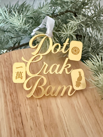 A gold metallic mahjong Christmas ornament spelling out 'Dot Crak Bam' with traditional mahjong tiles, hanging from a white ribbon on a wooden surface with green pine branches in the background.