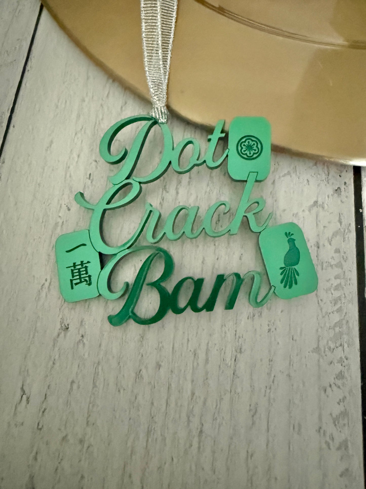 A green metallic mahjong Christmas ornament spelling out 'Dot Crak Bam' with traditional mahjong tiles, hanging from a silver ribbon on a wooden surface.