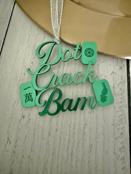 A green metallic mahjong Christmas ornament spelling out 'Dot Crak Bam' with traditional mahjong tiles, hanging from a silver ribbon on a wooden surface.