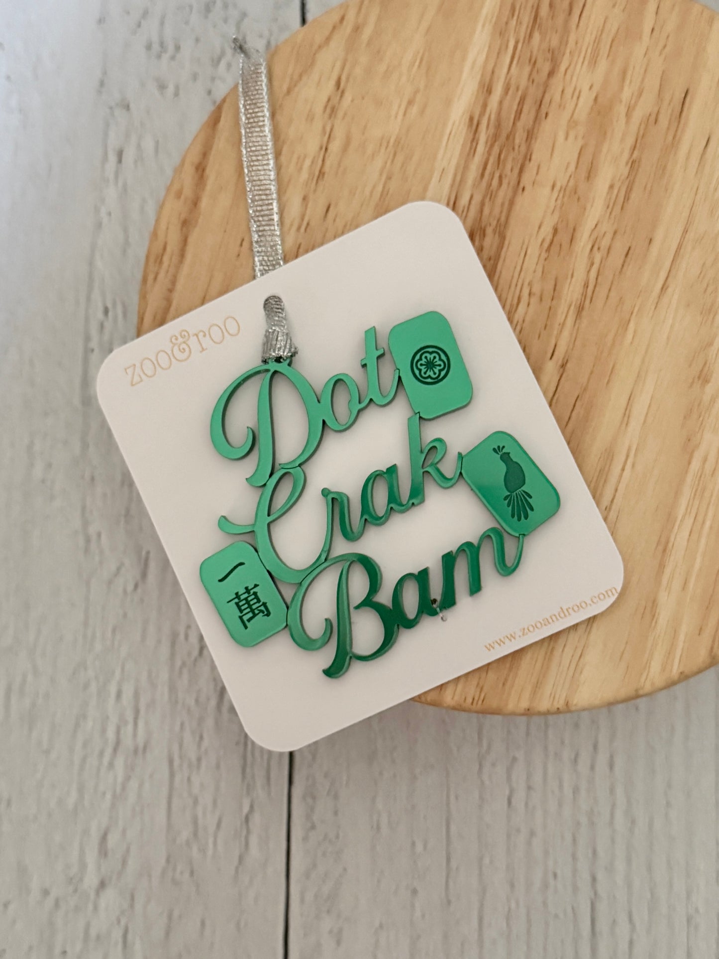 A green metallic mahjong ornament with the phrase “Dot Crak Bam” and mahjong tile graphics, attached to a white backing card with zoo&roo branding. The card is photographed on a round wooden board against a light wood plank background.