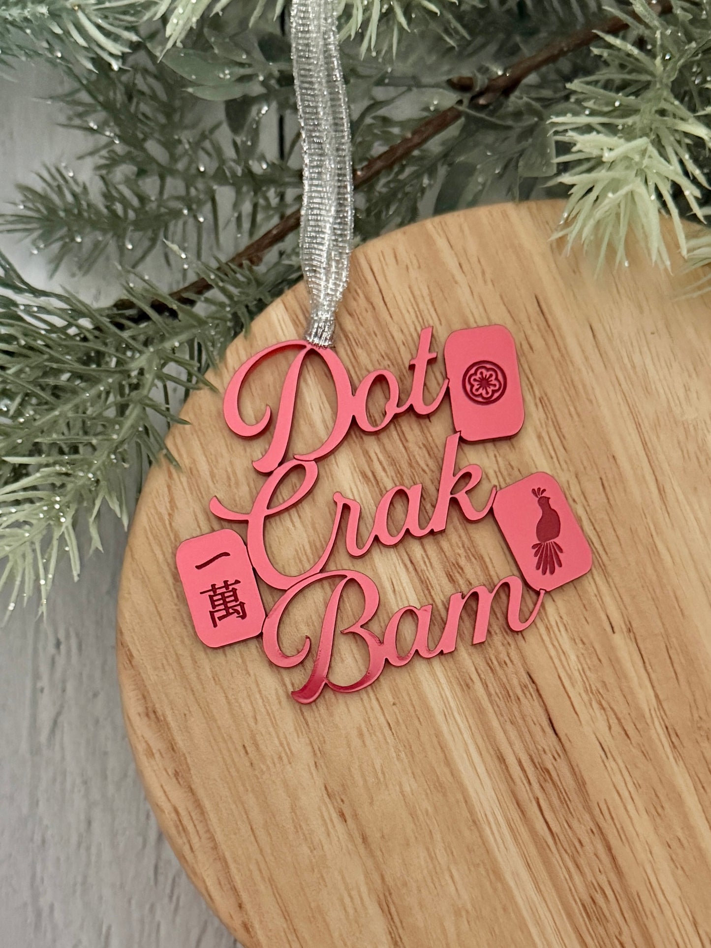 A red metallic mahjong Christmas ornament spelling out 'Dot Crak Bam' with traditional mahjong tiles, hanging from a silver ribbon on a wooden surface with green pine branches in the background.