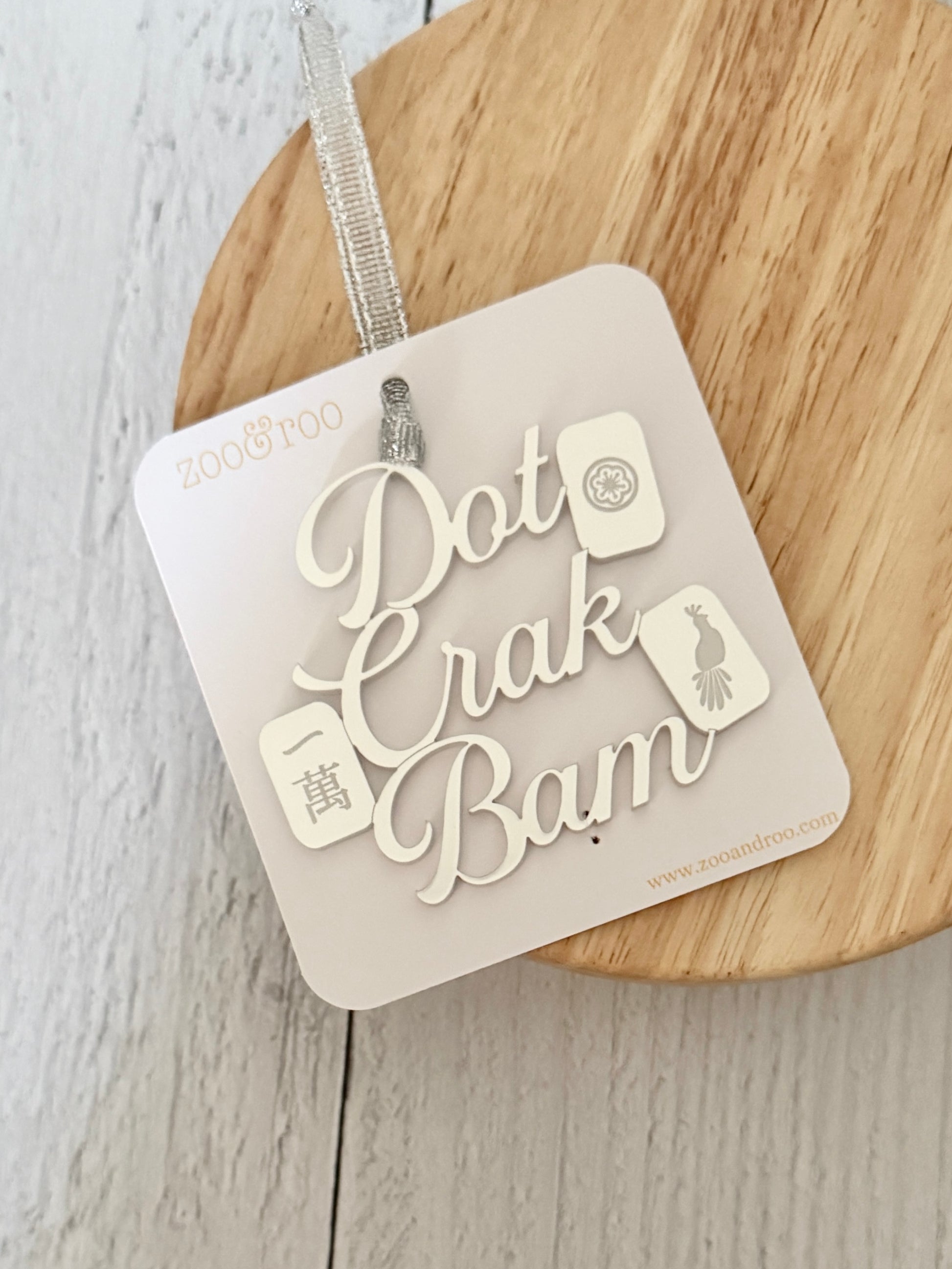 A silver metallic mahjong Christmas ornament spelling out 'Dot Crak Bam' with traditional mahjong tiles, hanging from a silver ribbon and attached to a white card, on a wooden surface.
