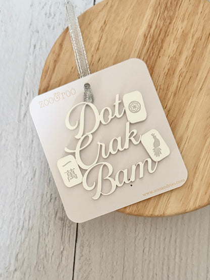 A silver metallic mahjong Christmas ornament spelling out 'Dot Crak Bam' with traditional mahjong tiles, hanging from a silver ribbon and attached to a white card, on a wooden surface.