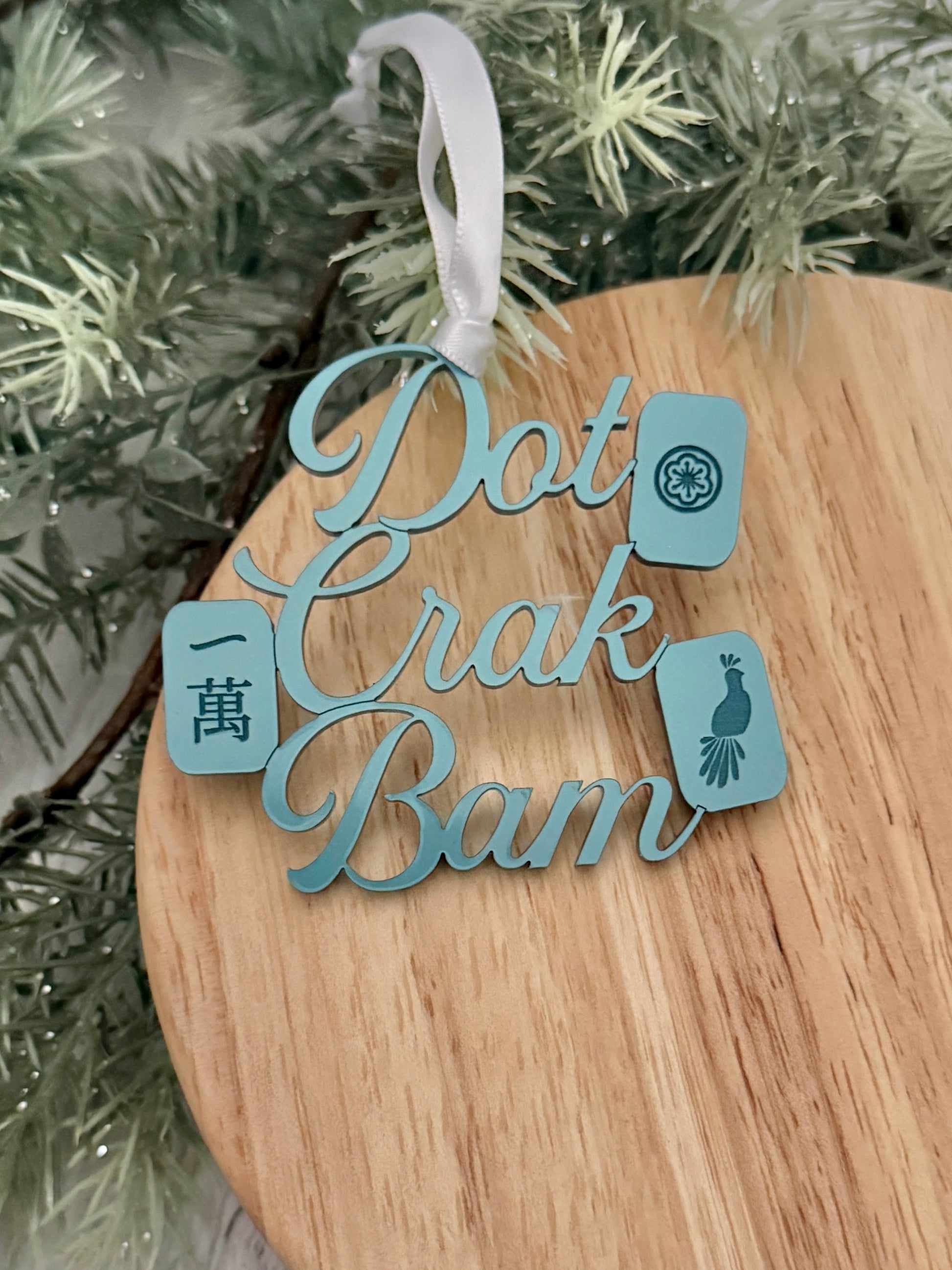 A turquoise metallic mahjong Christmas ornament spelling out 'Dot Crak Bam' with traditional mahjong tiles, hanging from a white ribbon on a wooden surface with green pine branches in the background.