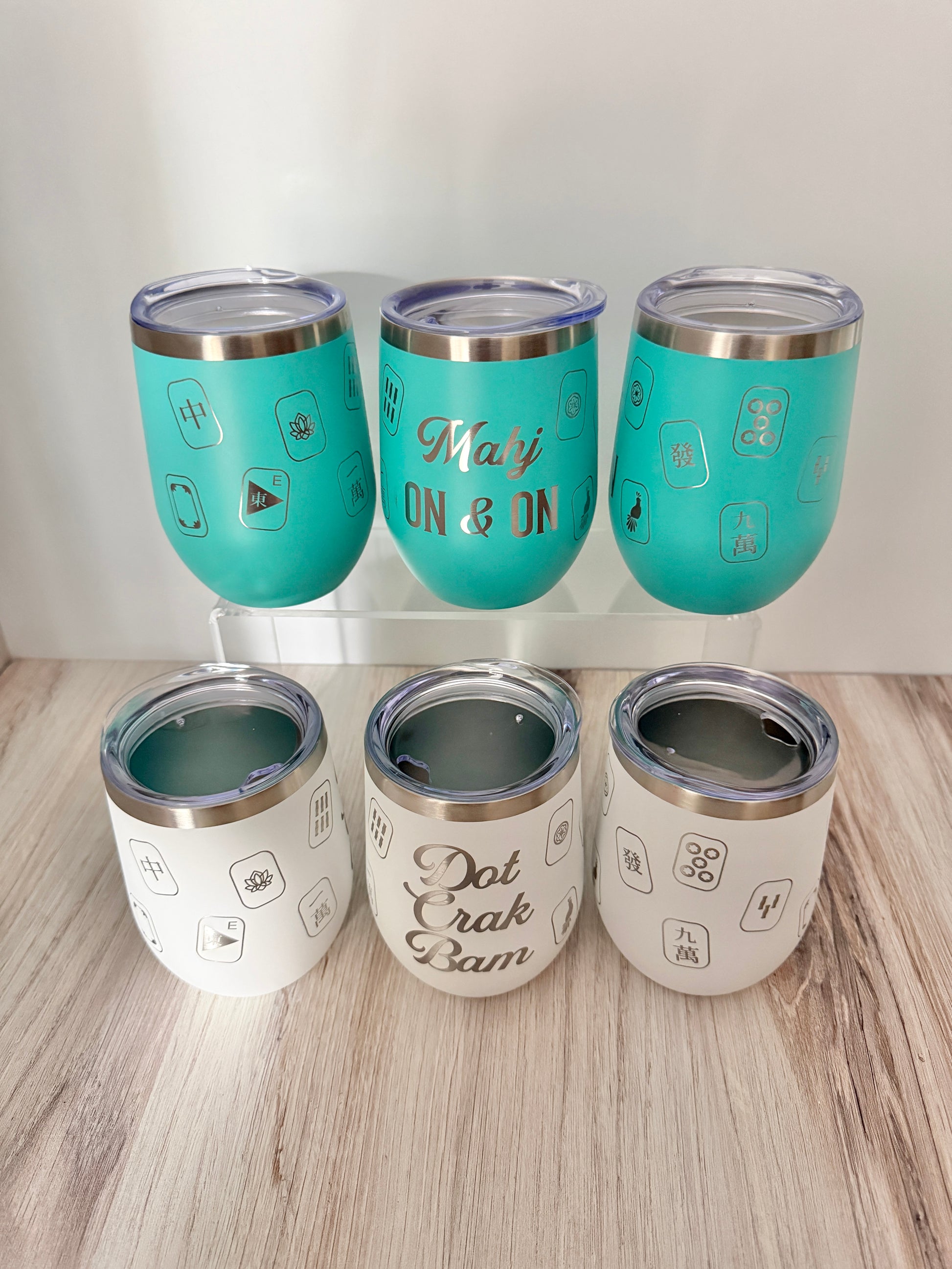 Six insulated stainless steel mahjong wine tumblers with clear lids are arranged on a light wood surface against a white wall. Three tumblers are turquoise with the text 'Mahj ON & ON'; the other three are white with engraved text 'Dot Crak Bam'.