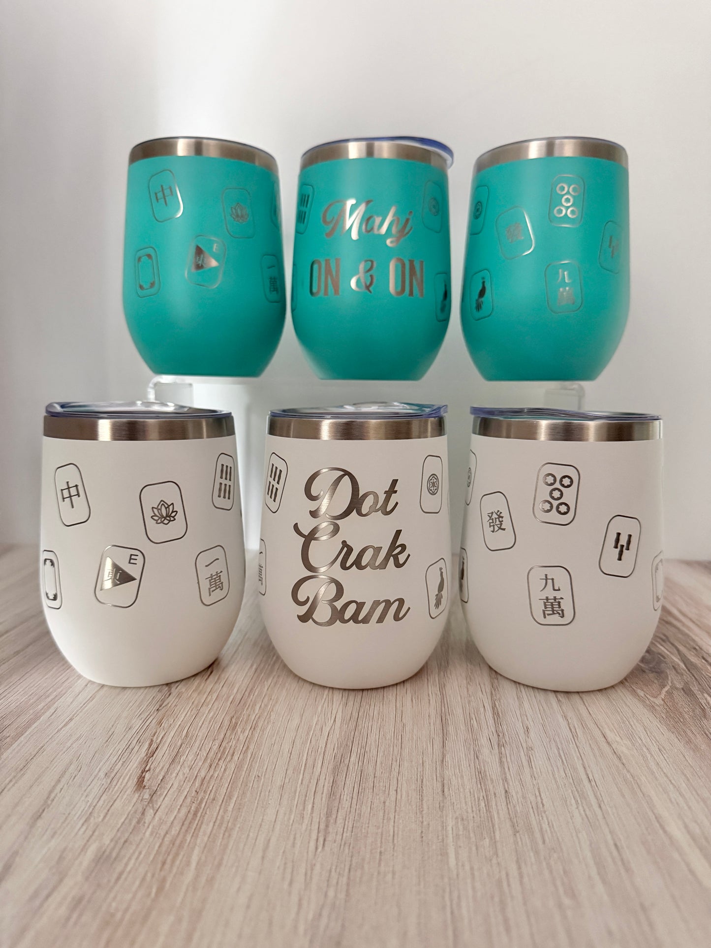 Six insulated stemless mahjong wine tumblers arranged on a light wood surface against a white wall. Three tumblers are turquoise with 'mahj on & on' text, and three are white with 'dot crak bam' text.