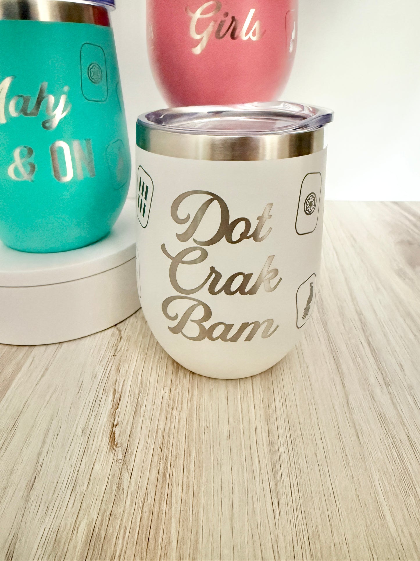 A white insulated stemless wine tumbler with a stainless steel rim and lid, featuring the phrase 'Dot Crak Bam' engraved. It is shown on a light wood surface next to other custom mahjong tumbler designs.