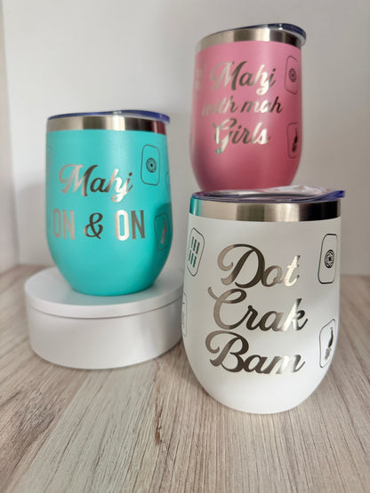 Three mahjong stainless steel insulated tumblers with clear lids are displayed on a light wood surface against a white wall. One tumbler is turquoise with engraved text reading 'Mahj ON & ON', one is pink with engraved text reading 'Mahj with mah girls', and one is white with engraved text reading 'Dot Crak Bam'.