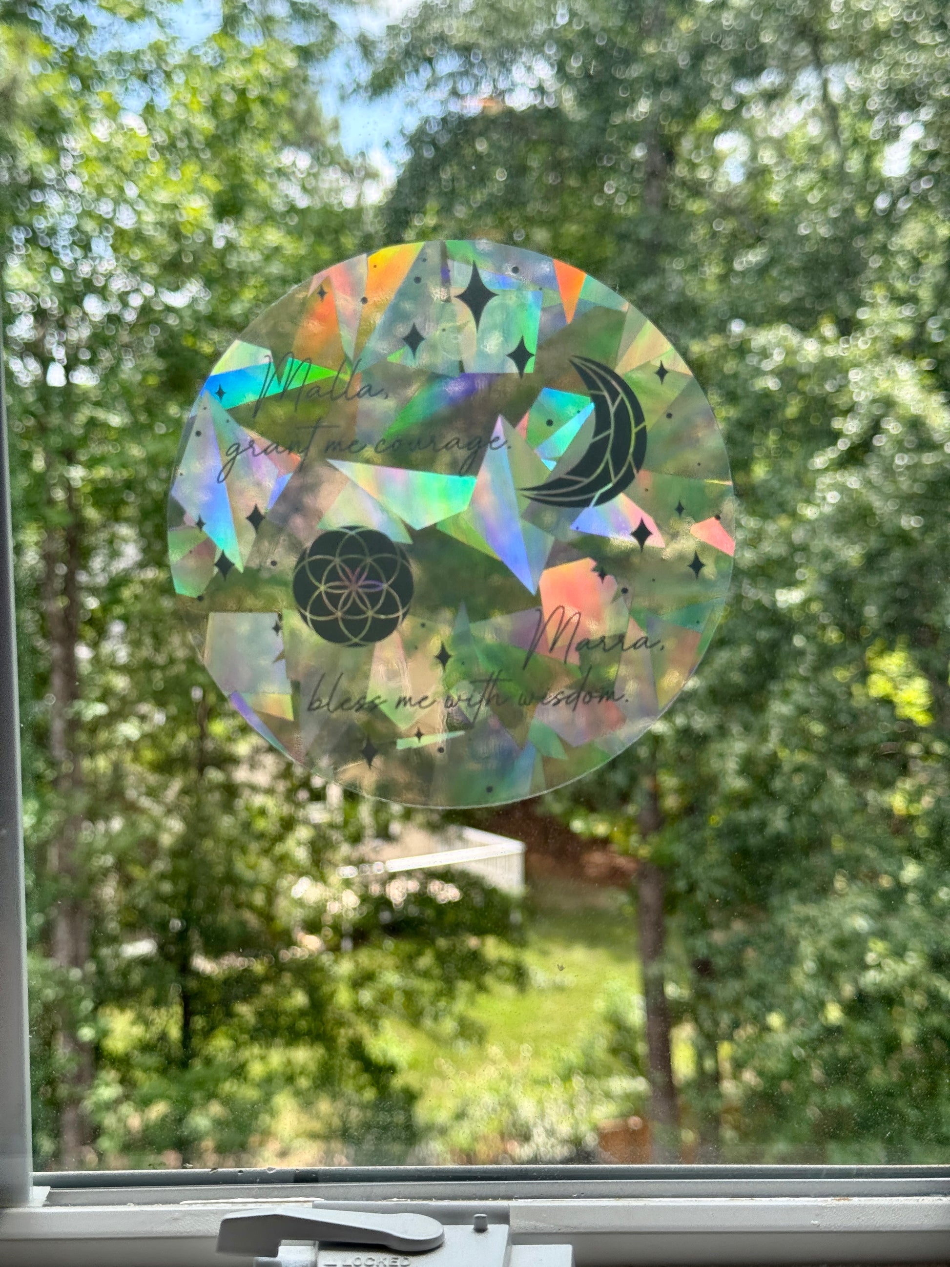 Decorative circular suncatcher inspired by The Road of Bones by Demi Winters with text 'Malla grant me courage. Marra bless me with wisdom' and moon and stars symbols on a window with a blurred outdoor background.